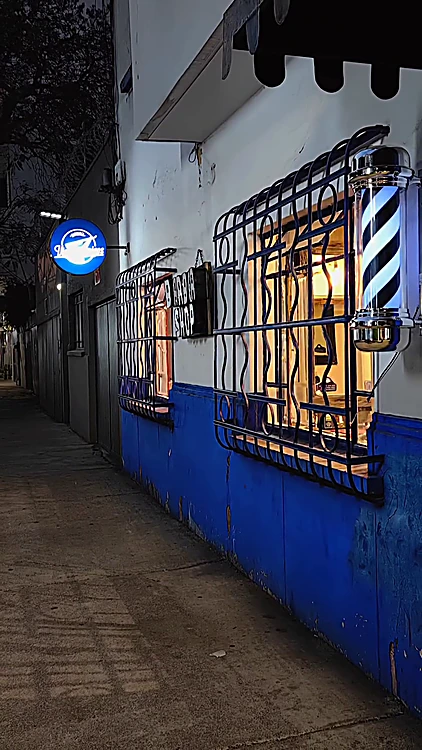 Fachada de noche: muro blanco con guardapolvo azul, ventana con herrería ondulada, poste de barbero y letrero redondo azul