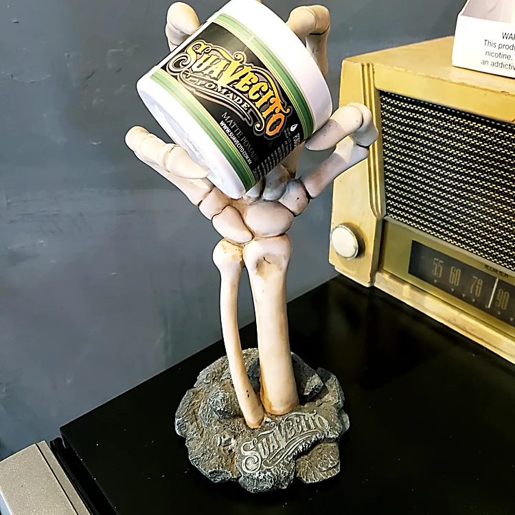 Figura de mano de esqueleto sosteniendo una lata de pomada Suavecito