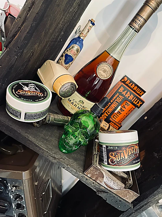 Repisa de madera con pomadas Suavecito, una botella con forma de calavera verde y una máquina